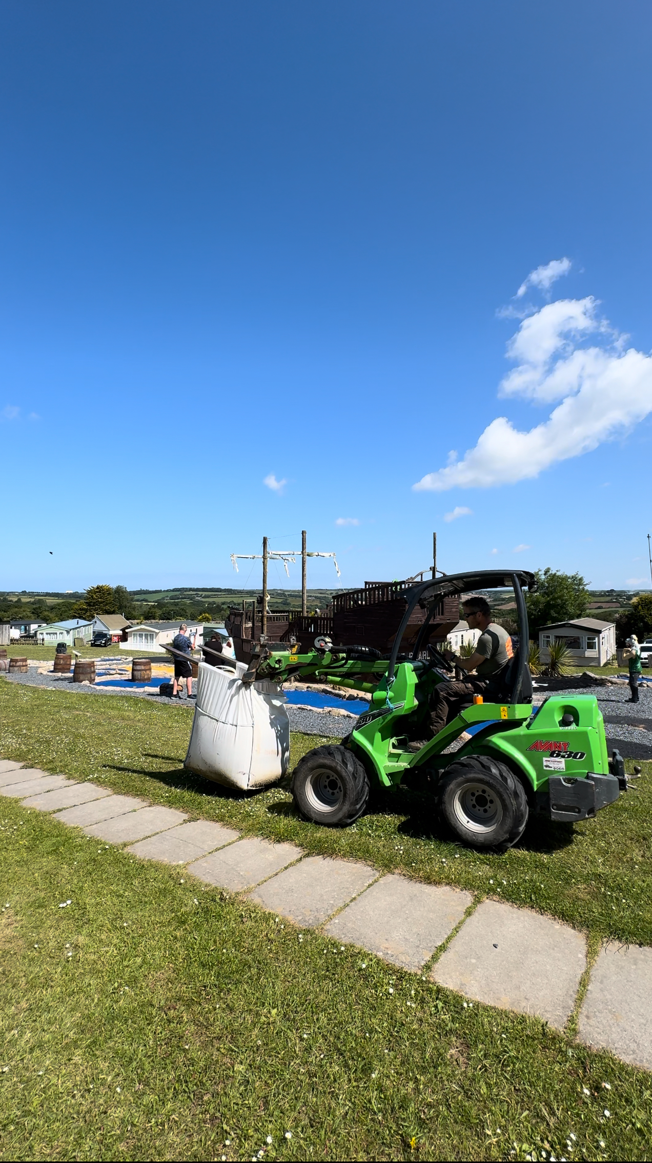 A compact loader carrying a bulk bag across a holiday park lawn